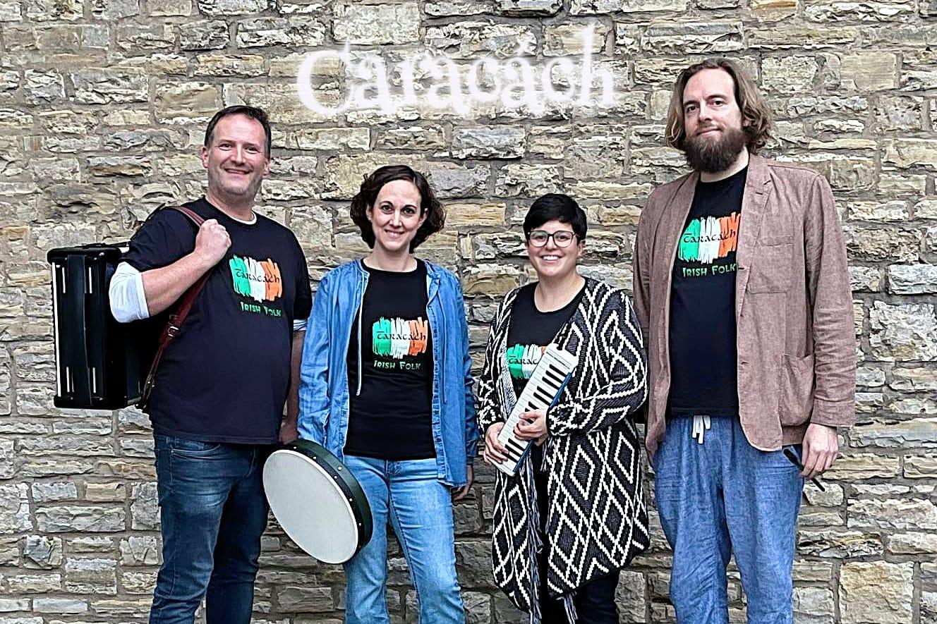 Copilot said: Vier Personen der Irish-Folk-Band stehen mit Instrumenten vor einer Natursteinmauer mit dem Schriftzug „Caracách“.