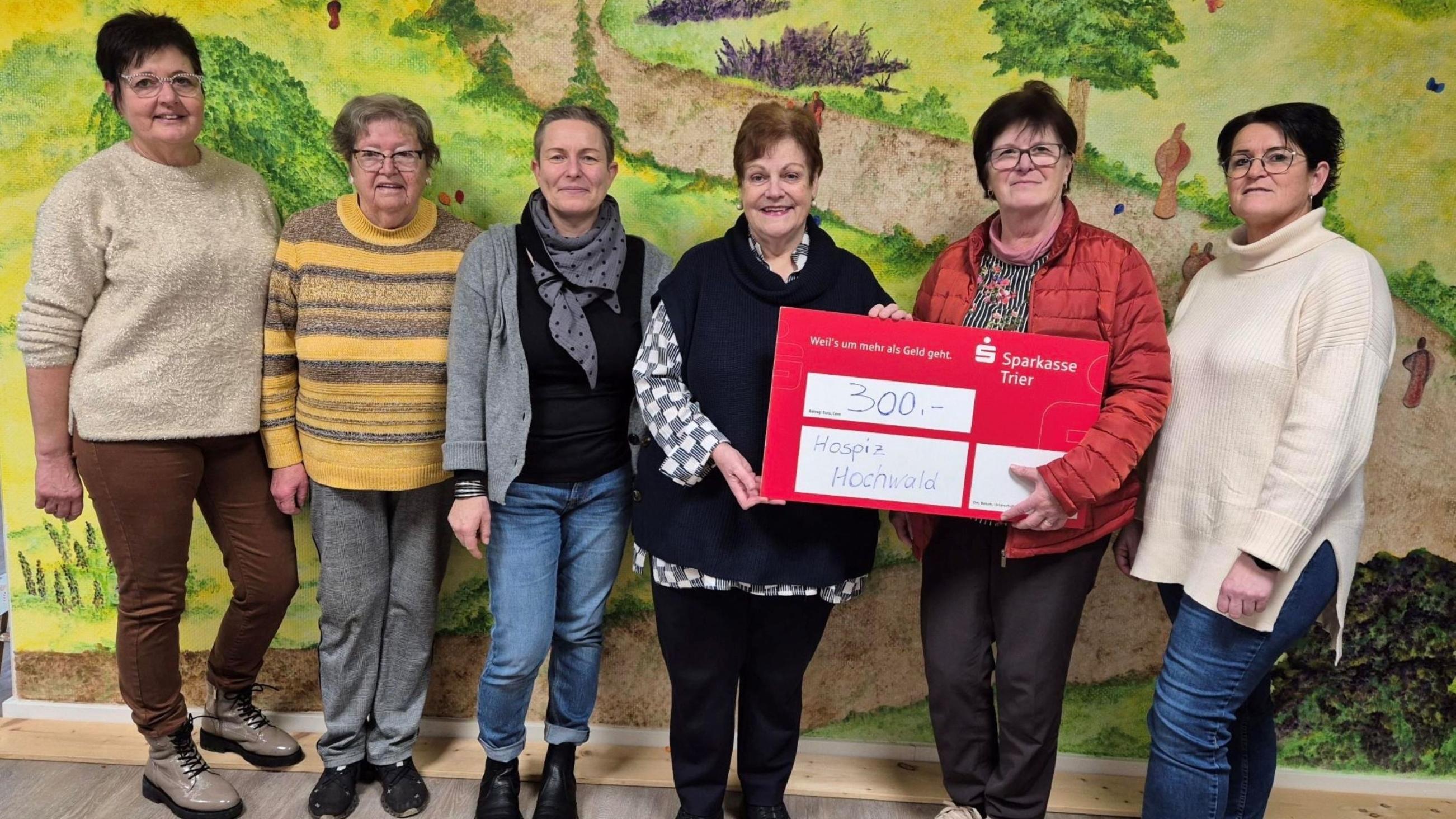 Sechs Personen stehen vor einer Wand mit einem bunten Landschaftsgemälde und halten gemeinsam einen großen roten Spendenscheck über 300 Euro für das „Hospiz Hochwald