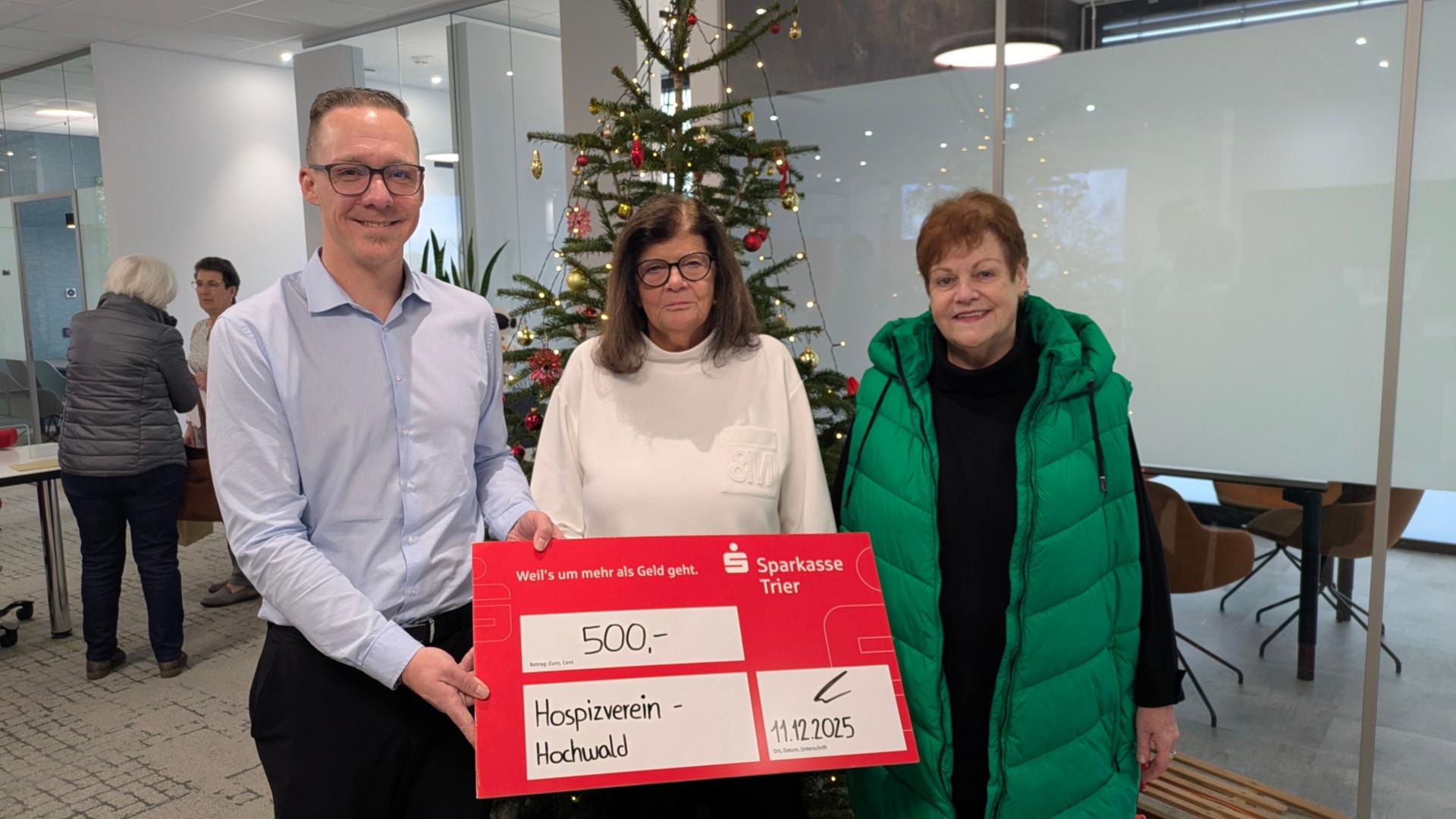 Das Foto zeigt eine Spendenübergabe in einem modernen Innenraum mit Weihnachtsdekoration. Im Vordergrund halten drei Personen einen großen symbolischen Scheck der Sparkasse Trier.