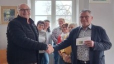 Zwei Personen stehen einander gegenüber und geben sich die Hand, während sie ein Dokument überreichen. Im Hintergrund steht eine kleine Gruppe.