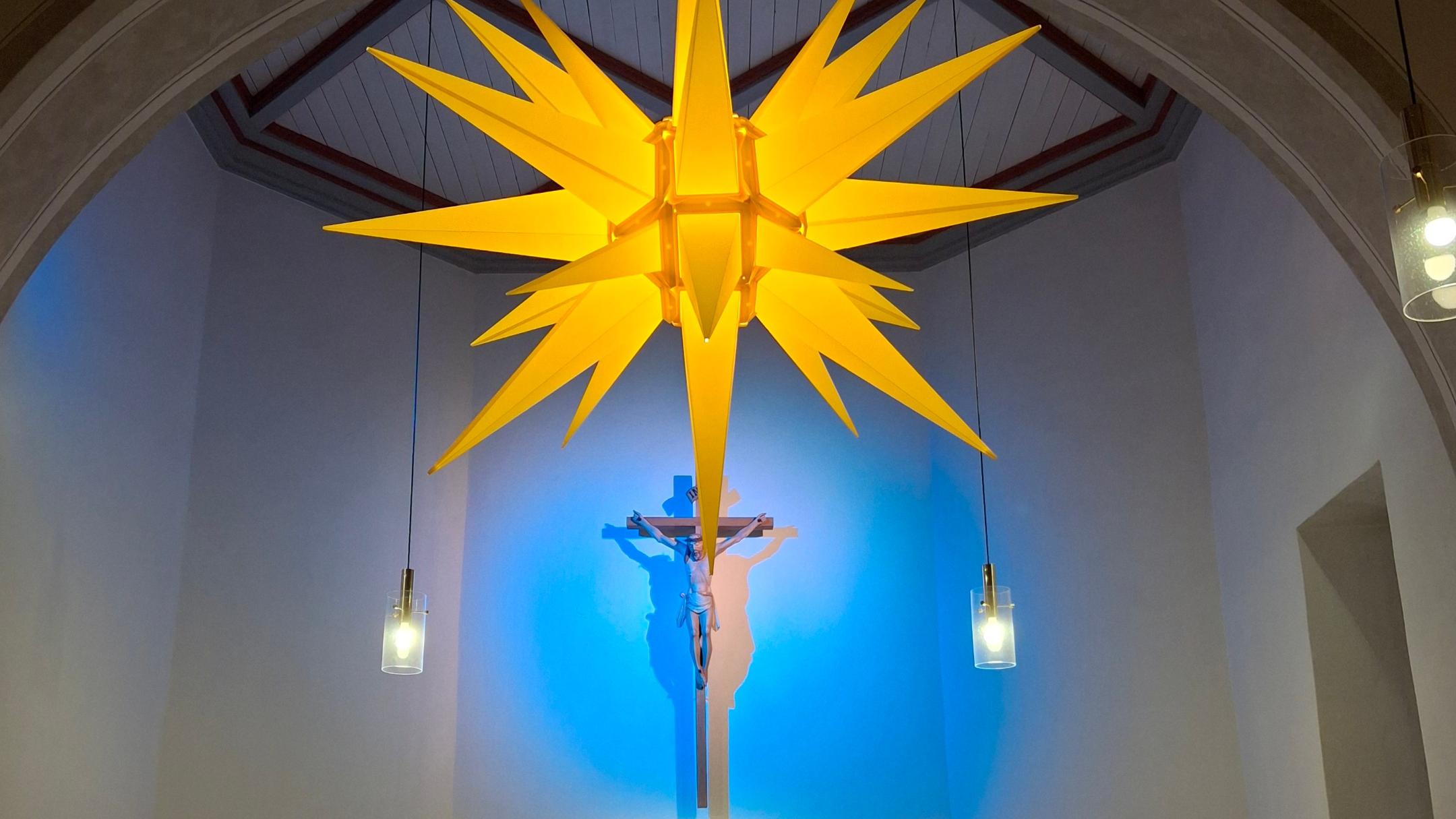 Das Bild zeigt einen hell erleuchteten Kirchenaltar mit einer großen gelben Weihnachtsstern-Laterne im Vordergrund, Kerzen und Blumen auf dem Altar sowie einem goldenen Tabernakel, während ein blau angestrahltes Kreuz im Hintergrund hängt.