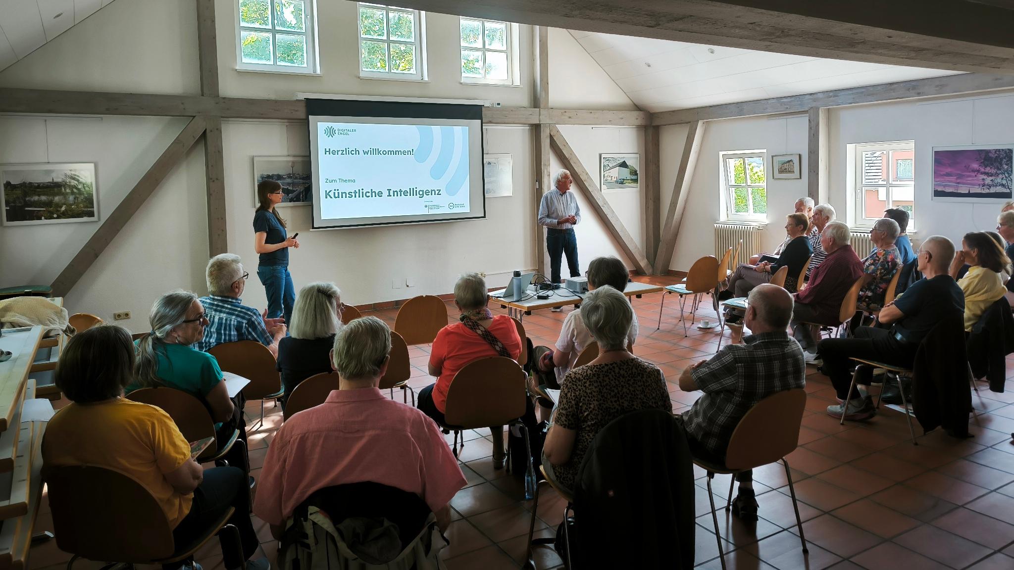 Teilnehmende verfolgen in einem hellen Saal mit Holzbalken aufmerksam einen Vortrag der Digitalen Engel zum Thema Künstliche Intelligenz.