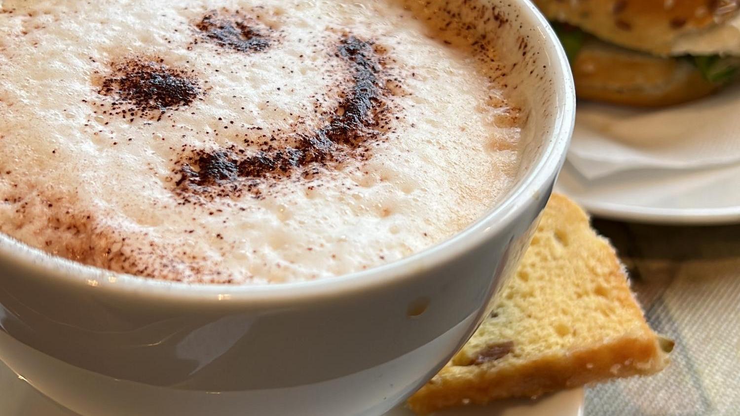 Eine Tasse Cappuccino mit Smiley aus Kakao steht neben einem Bagel und einem Stück Kuchen.