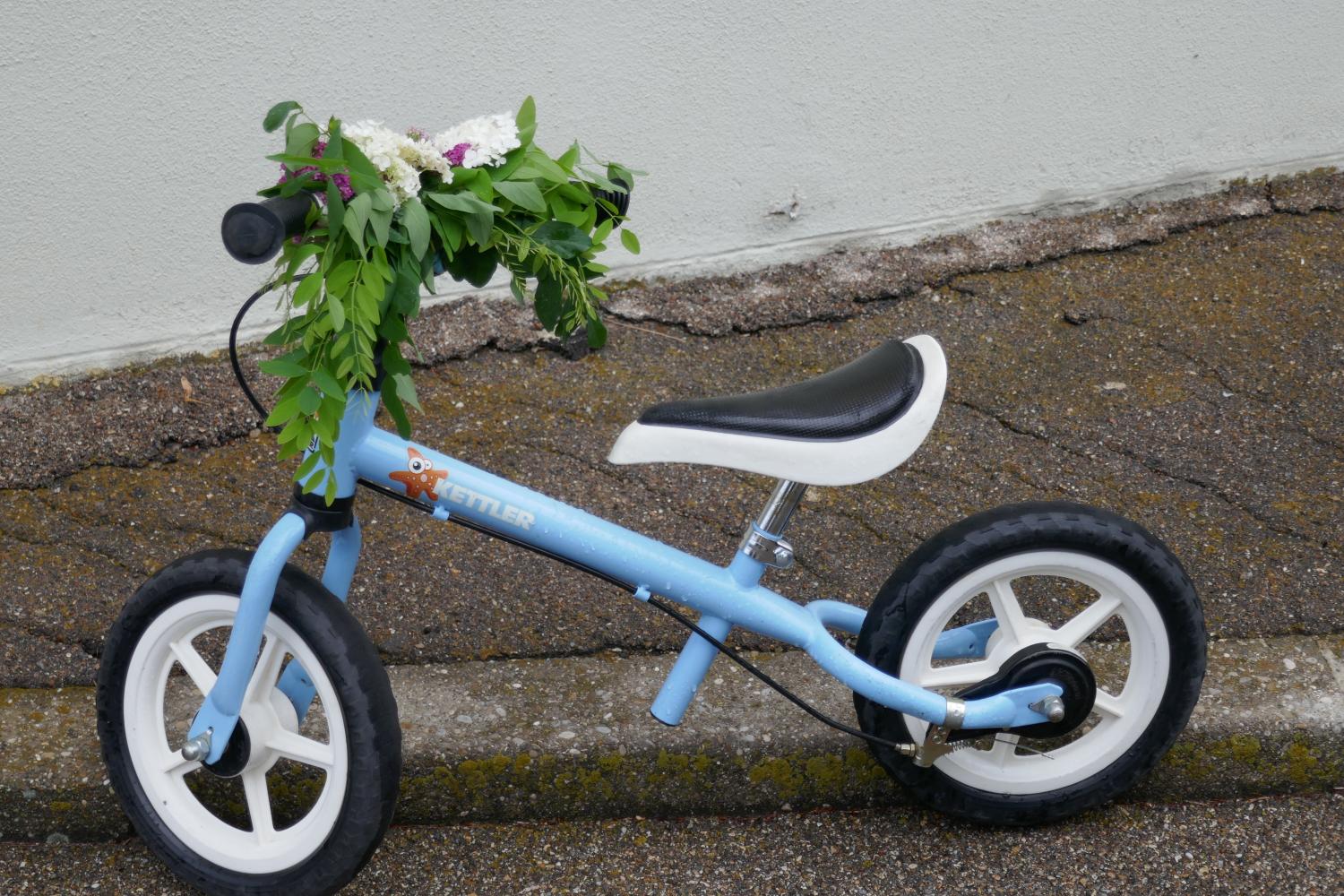 Blaues Kinderlaufrad mit Blumen am Lenker auf Gehweg abgestellt.