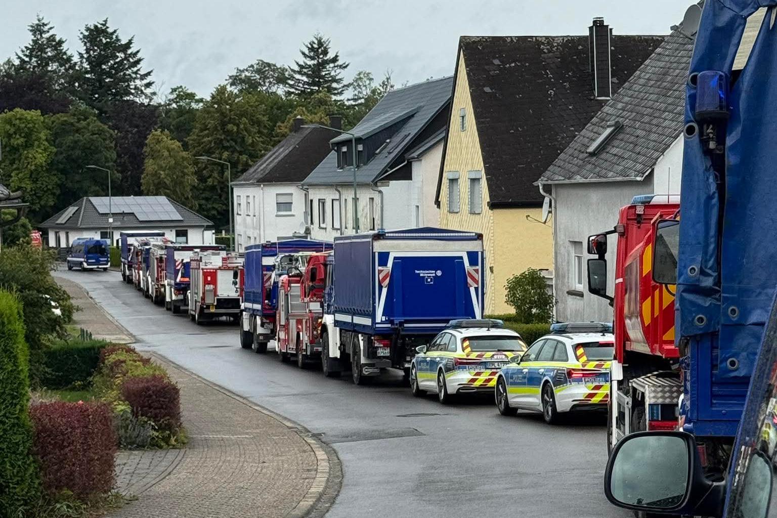 Nasse Wohnstraße mit einer langen Reihe von Feuerwehrfahrzeugen und Polizeiautos vor Wohnhäusern unter grauem Himmel.