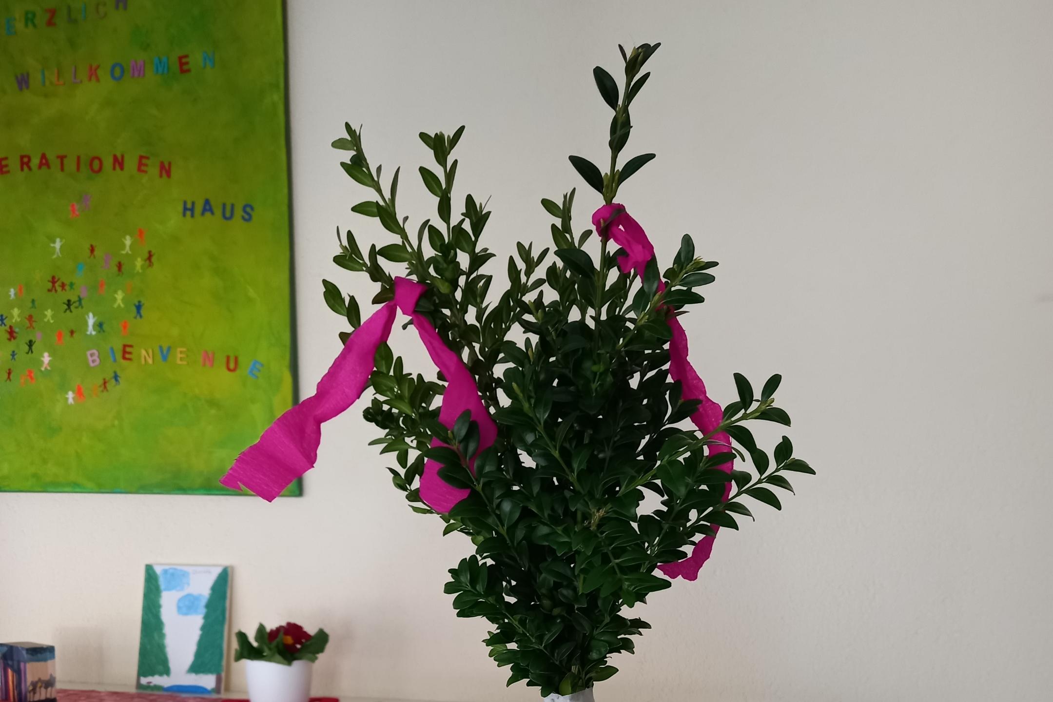 Ein kleiner geschmückter Strauch in einem weißen Topf steht auf einem Tisch mit einer grünen Filzunterlage.