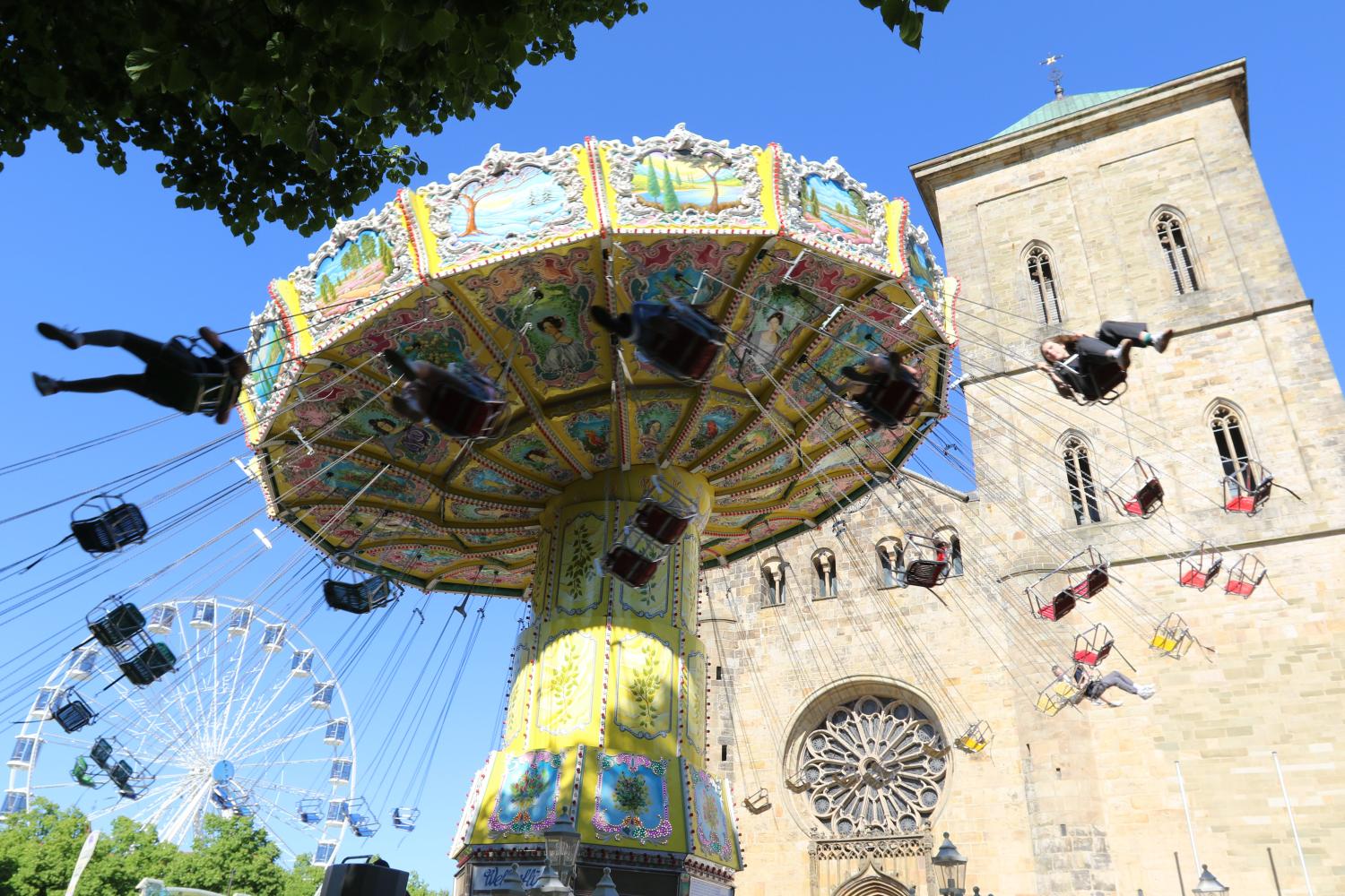 Kettenkarussell mit Menschen vor einer Kirche bei sonnigem Wetter, Riesenrad im Hintergrund.