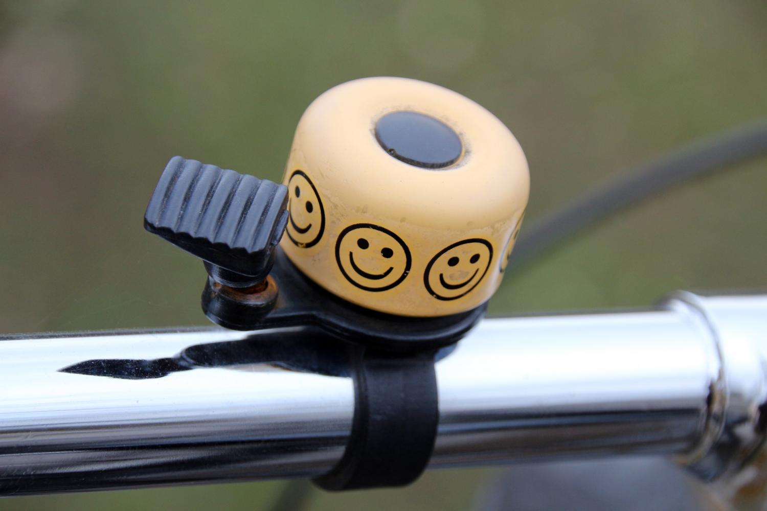 Eine gelbe Fahrradklingel mit mehreren aufgedruckten Smileys ist an einem silbernen Lenker befestigt.