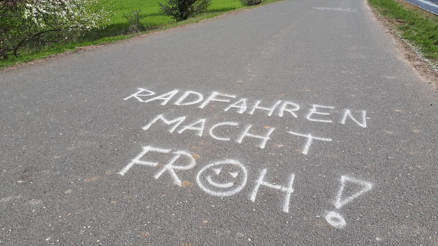 Text „Radfahren macht froh!“ auf asphaltiertem Radweg, blühende Bäume am Rand.
