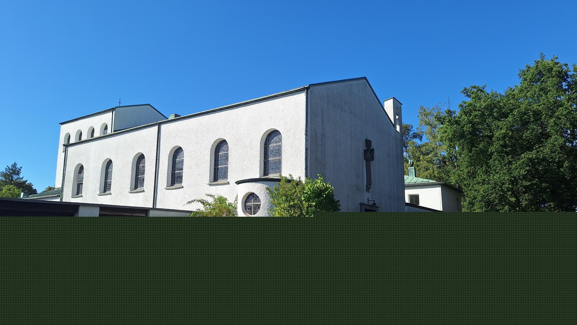 Das Bild zeigt eine weiße Kirche mit Rundbogenfenstern und einem Kreuz an der Fassade, sie steht umgeben von Bäumen und Grünflächen. Der Himmel ist klar und blau, was der Szene eine ruhige, friedliche Stimmung verleiht.