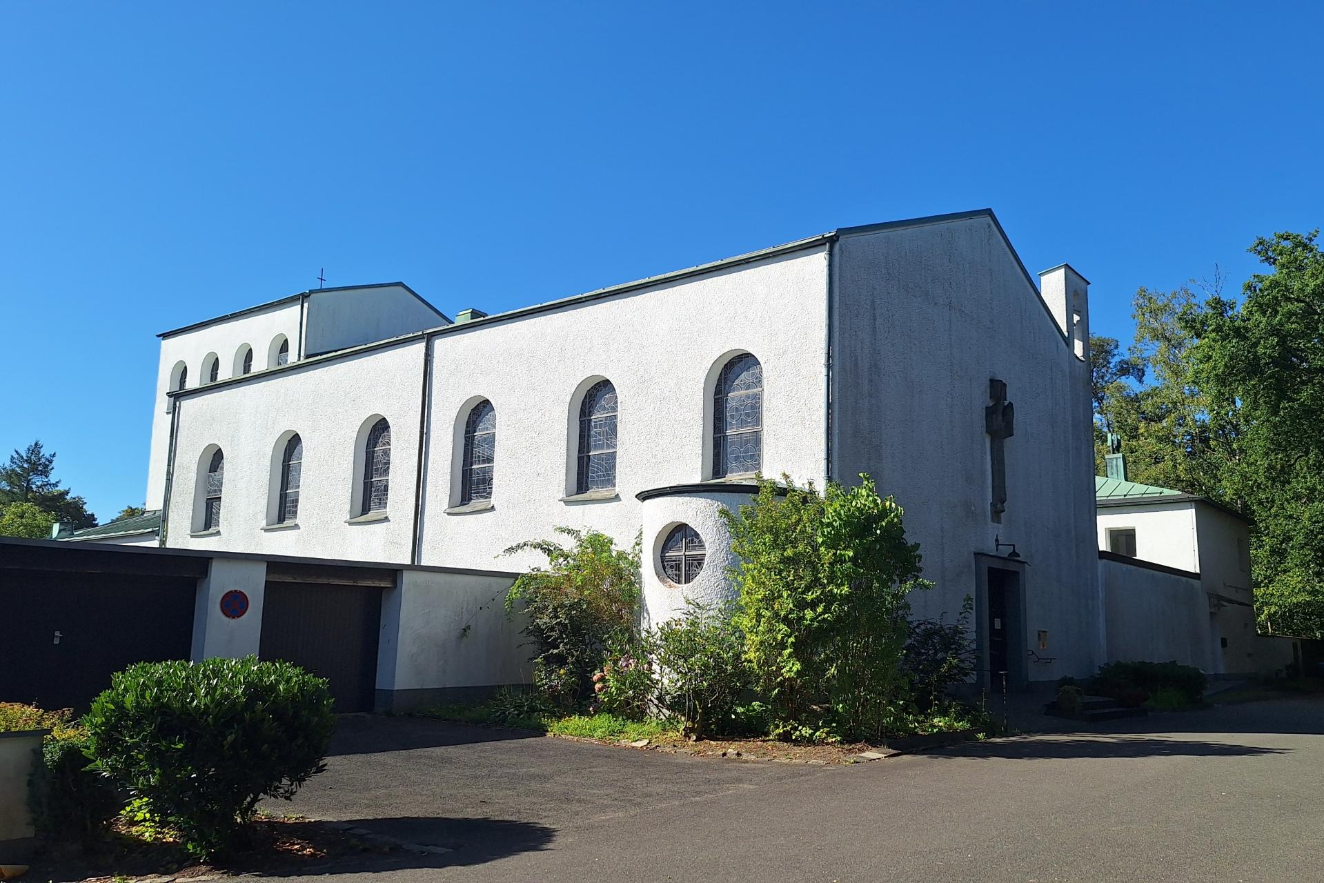Das Bild zeigt eine weiße Kirche mit Rundbogenfenstern und einem Kreuz an der Fassade, sie steht umgeben von Bäumen und Grünflächen. Der Himmel ist klar und blau, was der Szene eine ruhige, friedliche Stimmung verleiht.