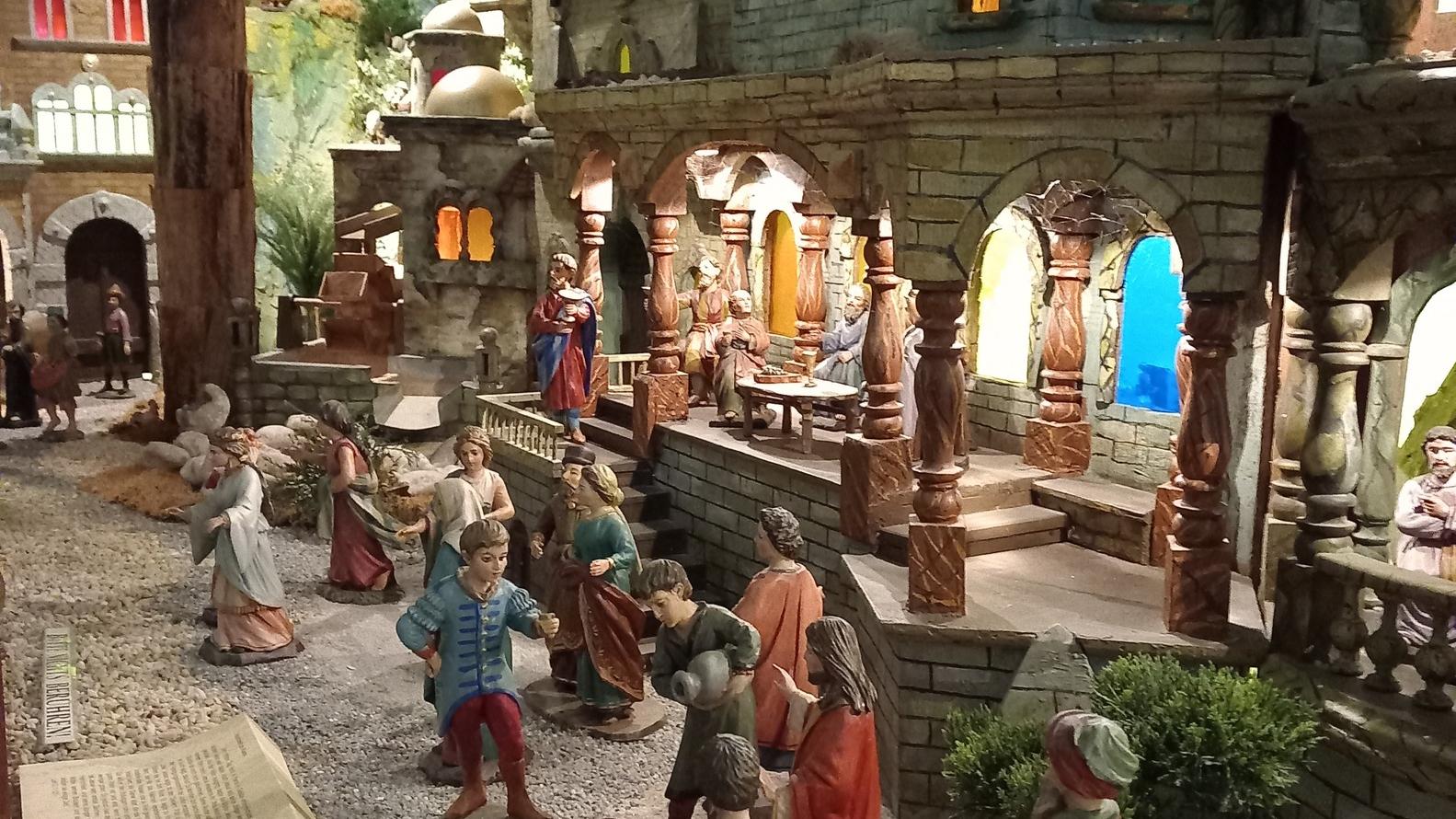 Die Szene zeigt eine detailreiche Weihnachtskrippe mit vielen handgeschnitzten Figuren, die auf einem steinigen Weg vor einem großen orientalisch anmutenden Gebäude stehen, während im Hintergrund weitere Figuren in beleuchteten Arkaden und Häusern das Leben zur Zeit Jesu darstellen.